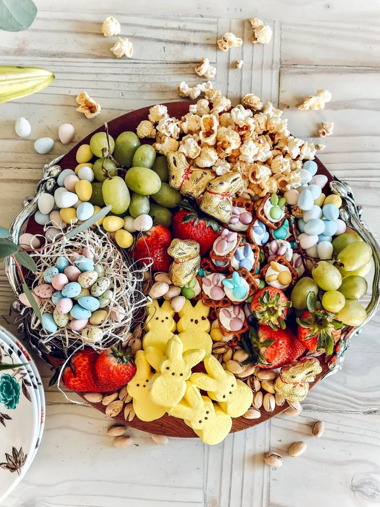 Easter dessert charcuterie board featuring yellow Peeps marshmallow bunnies, chocolate gold bunnies, pastel candy eggs in straw nest, popcorn, yogurt-covered pretzels, strawberries, grapes, and pistachios on a wooden platter