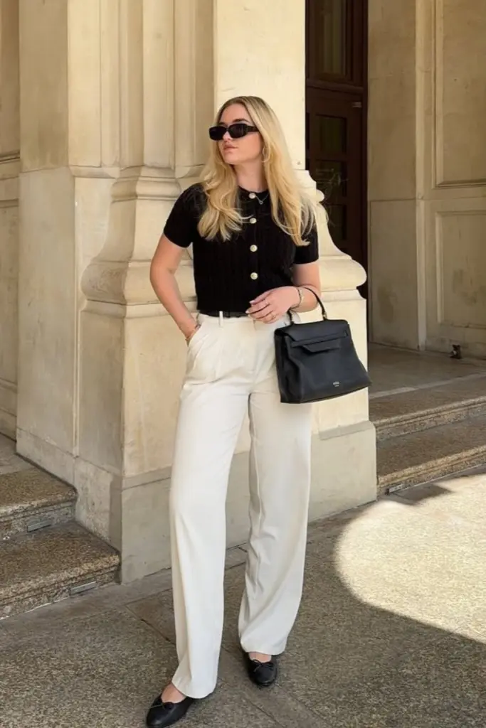 Business casual outfit with black short-sleeve knit cardigan with gold buttons, white wide-leg trousers, black flats and black structured handbag
