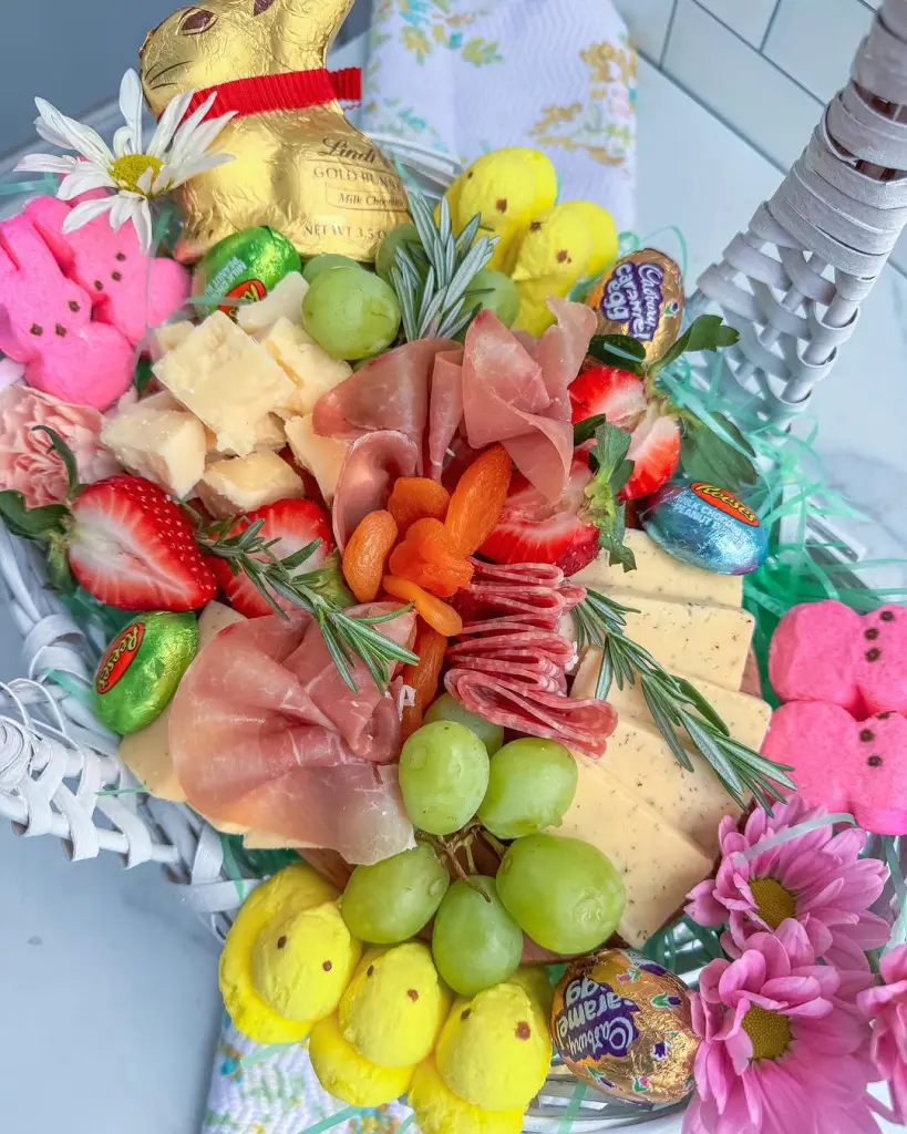 White Easter basket charcuterie featuring pink Peeps, Lindt gold bunny, Cadbury eggs, prosciutto, salami, cheese cubes, strawberries, grapes, dried apricots, and fresh daisies on decorative spring napkin
