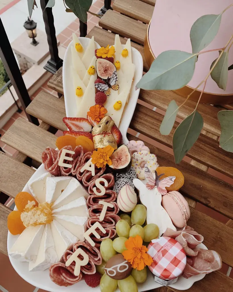Easter charcuterie board featuring cheese, salami letters spelling "EASTER," chocolate bunny, fresh figs, macarons, and spring florals on a white platter.
