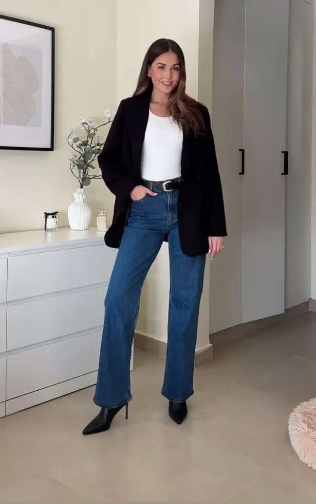 Business casual outfit with oversized black blazer, white bodysuit, wide-leg blue jeans, black belt and pointed ankle boots in minimalist home setting