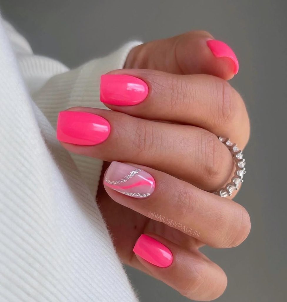Short squoval nails with bright neon pink polish and clear base with pink and silver glitter wave design on ring finger 