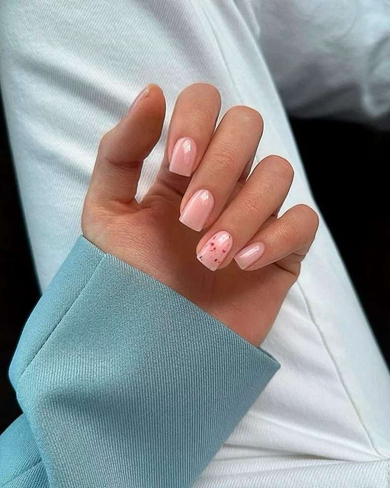 Short square nails with sheer pink polish and one accent nail featuring small red speckles