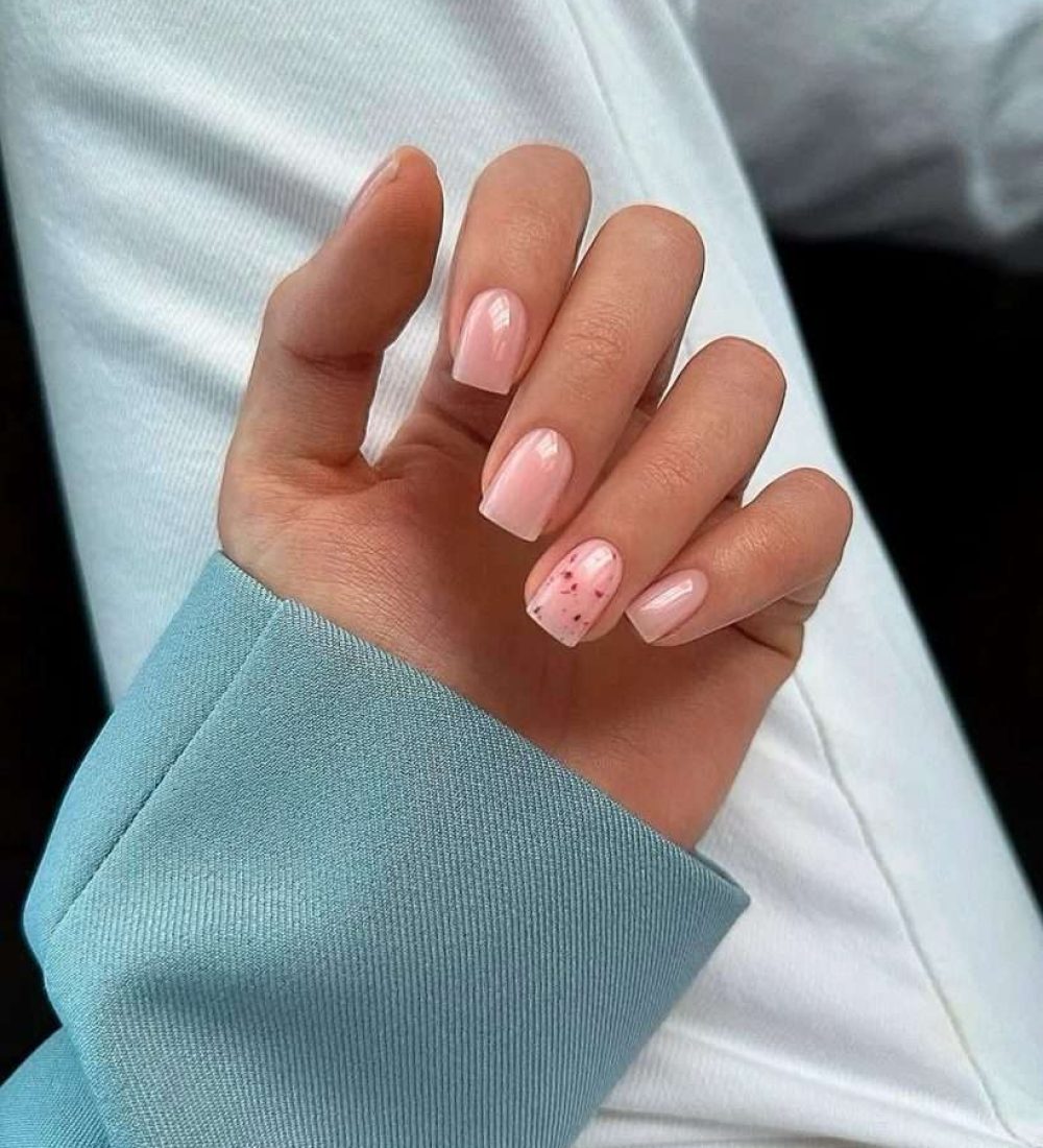 : Short square nails with sheer pink polish and one accent nail featuring small red speckles