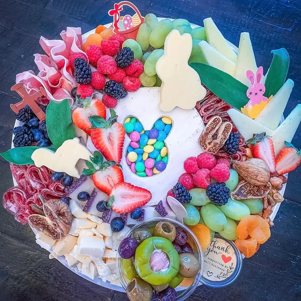 Easter charcuterie board with bunny-shaped cheese cutouts, candy-filled egg centerpiece, fresh berries, charcuterie, grapes, and colorful accents on a white platter with decorative Easter picks