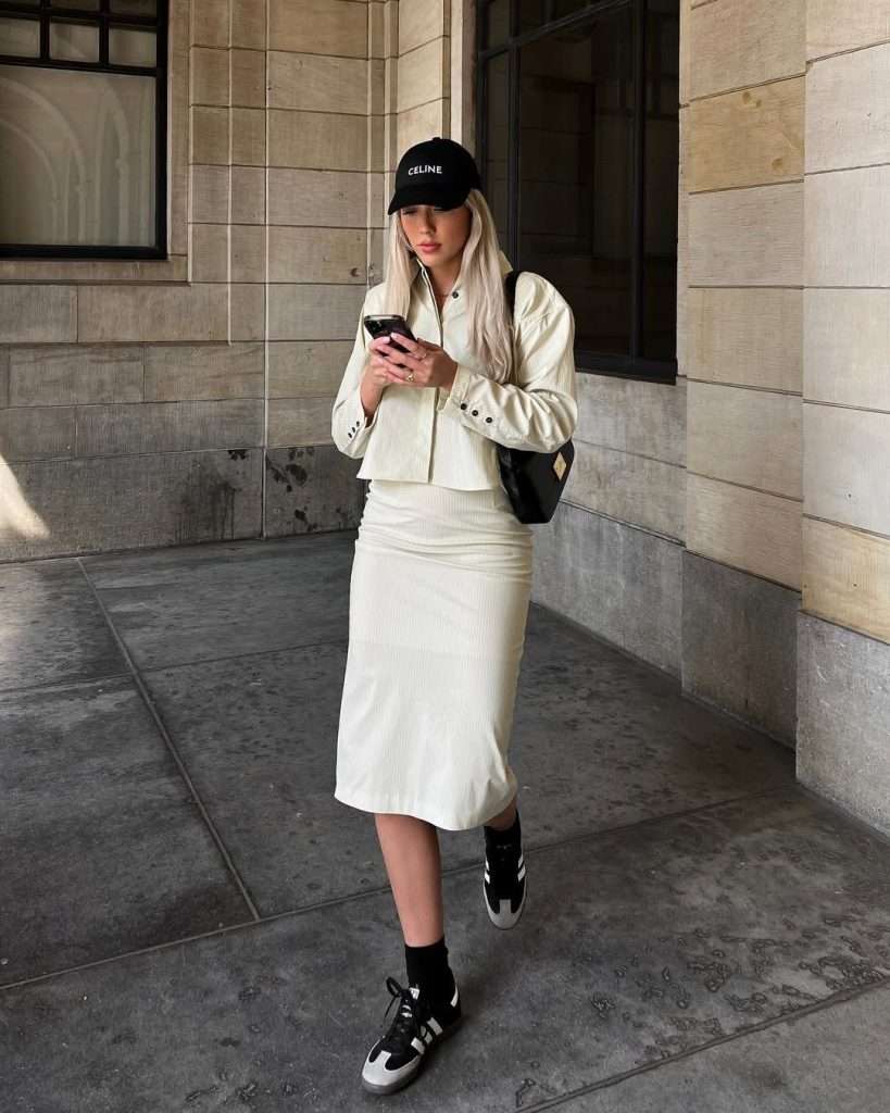 casual spring outfit with white cropped jacket, white ribbed midi skirt, black Celine baseball cap, black socks and black and gray Adidas sneakers