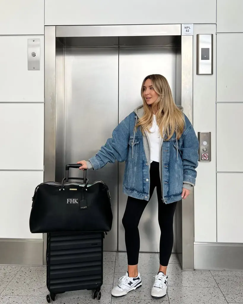 Casual airport outfit with oversized denim jacket, gray hoodie, black leggings, white sneakers and black monogrammed weekender bag with rolling suitcase