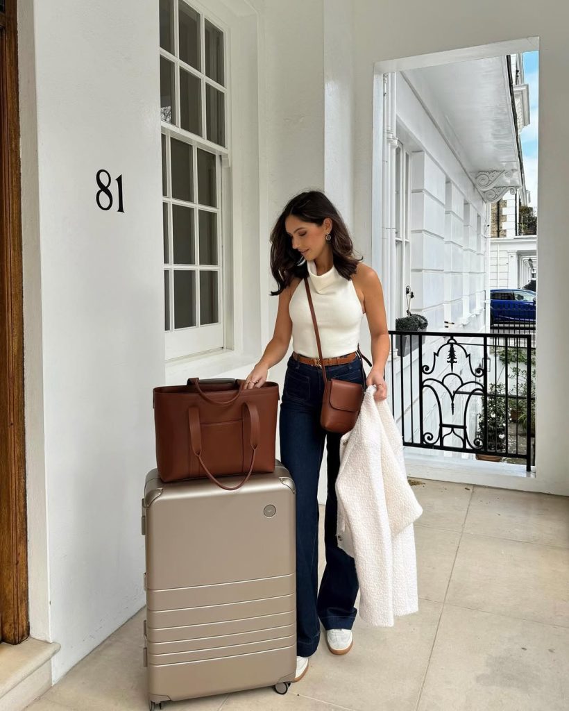 Elegant travel outfit with white halter neck top, dark flare jeans, brown belt, brown crossbody bag, white sneakers and cream coat with brown leather tote

