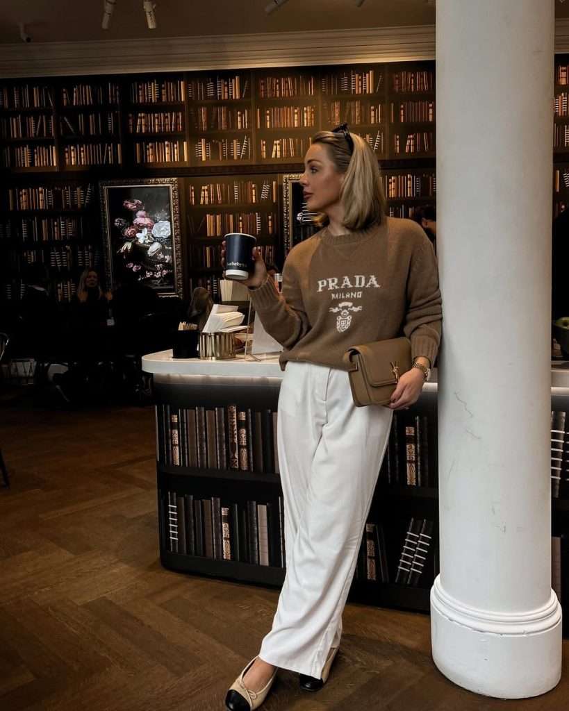 classy spring outfit with brown logo sweater, white wide-leg trousers, black and beige ballet flats and tan shoulder bag in a library setting
