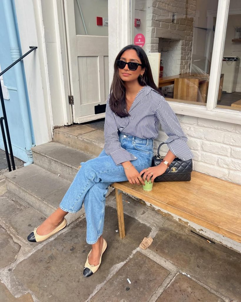 Summer business casual outfit with blue and white striped button-down shirt, light blue straight-leg jeans, beige and black cap-toe ballet flats, quilted black handbag and black sunglasses