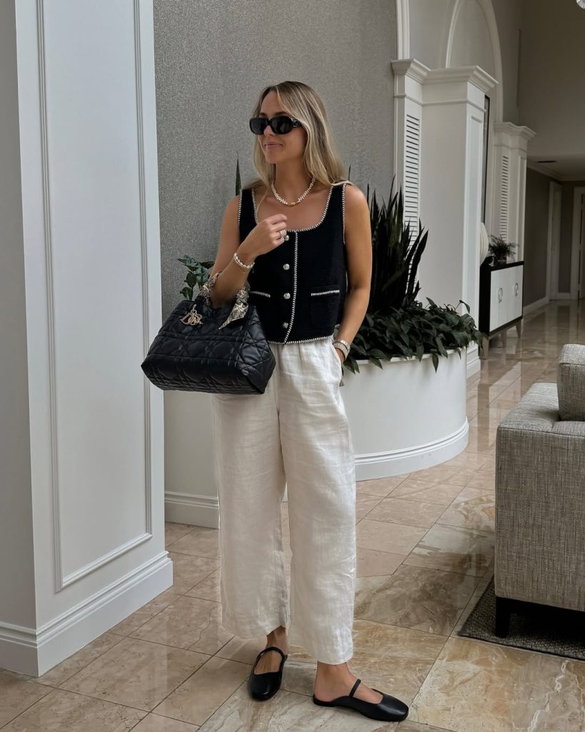 Elegant monochrome outfit with black contrast-trim vest, white wide-leg linen pants, pearl necklace, quilted black handbag and black mary jane flats
