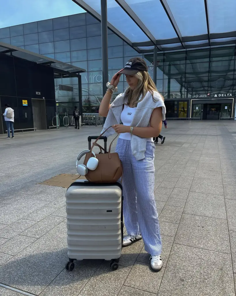 Comfortable airport outfit with white tank top, blue striped wide-leg pants, gray hoodie, black baseball cap, white sneakers and tan leather tote bag