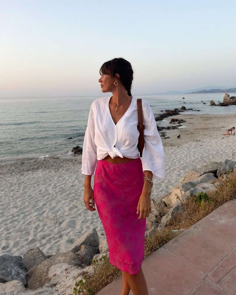 Beach vacation outfit with white linen button-down shirt tied at waist, bright pink textured midi skirt, gold hoop earrings, pendant necklace and brown leather crossbody bag