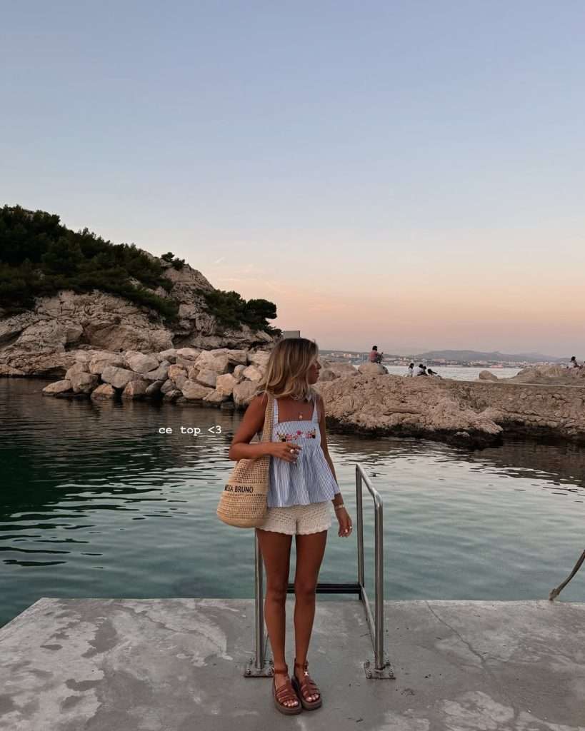 Beach vacation outfit with white and blue striped tank top featuring floral embroidery, cream crochet shorts, designer straw bucket bag and brown leather platform sandals