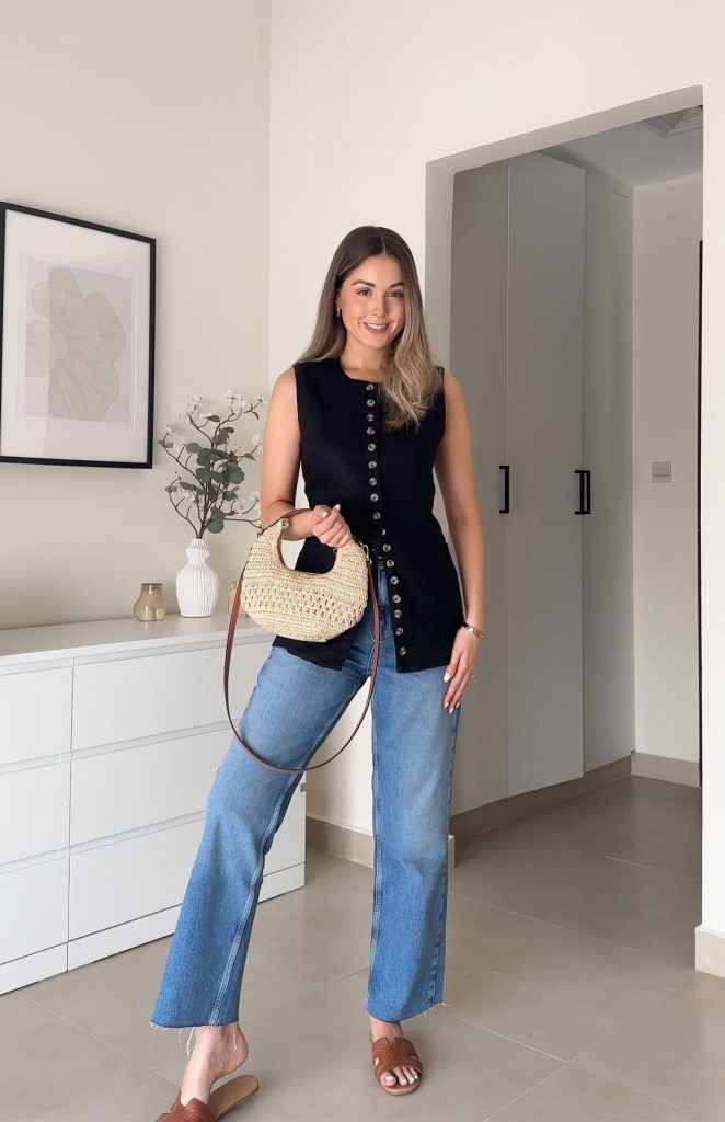 Summer business casual outfit with black sleeveless button-up vest, light blue wide-leg jeans, brown leather slide sandals and small woven straw crossbody bag with leather strap
