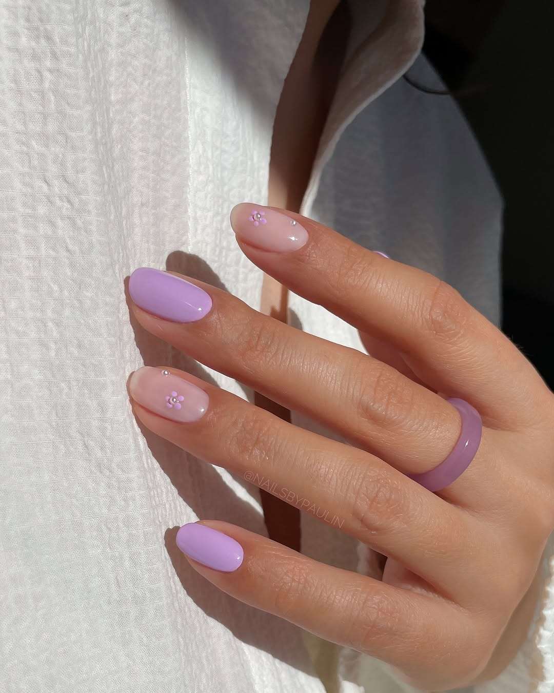 Short oval nails alternating between lavender purple and milky white with pearl flower accents for elegant spring manicure