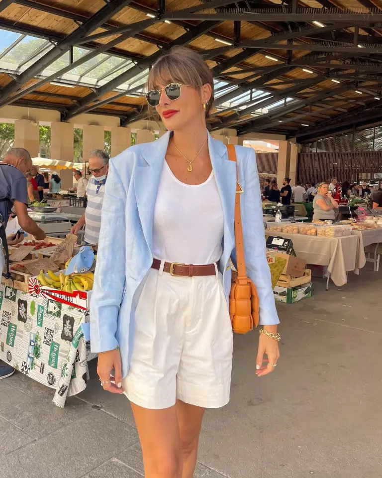 European vacation outfit with light blue blazer, white top, white tailored shorts, brown leather belt, matching loafers and tan crossbody bag