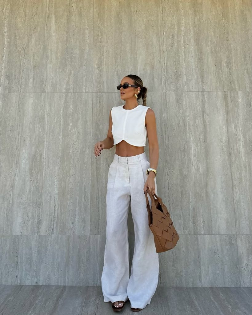 Elegant minimalist outfit with white structured crop top, white wide-leg linen pants, tan leather handbag, gold statement earrings and slim black sunglasses
