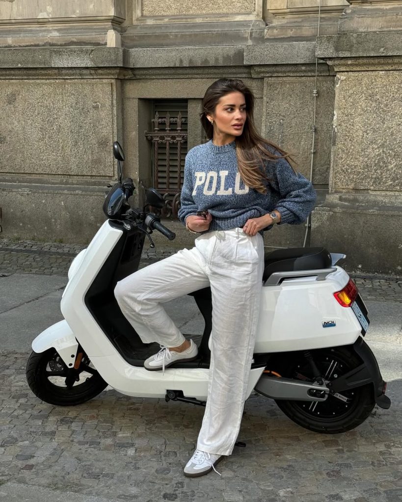 Casual chic outfit with blue collegiate logo sweater, white wide-leg linen pants, white sneakers and vintage scooter setting
