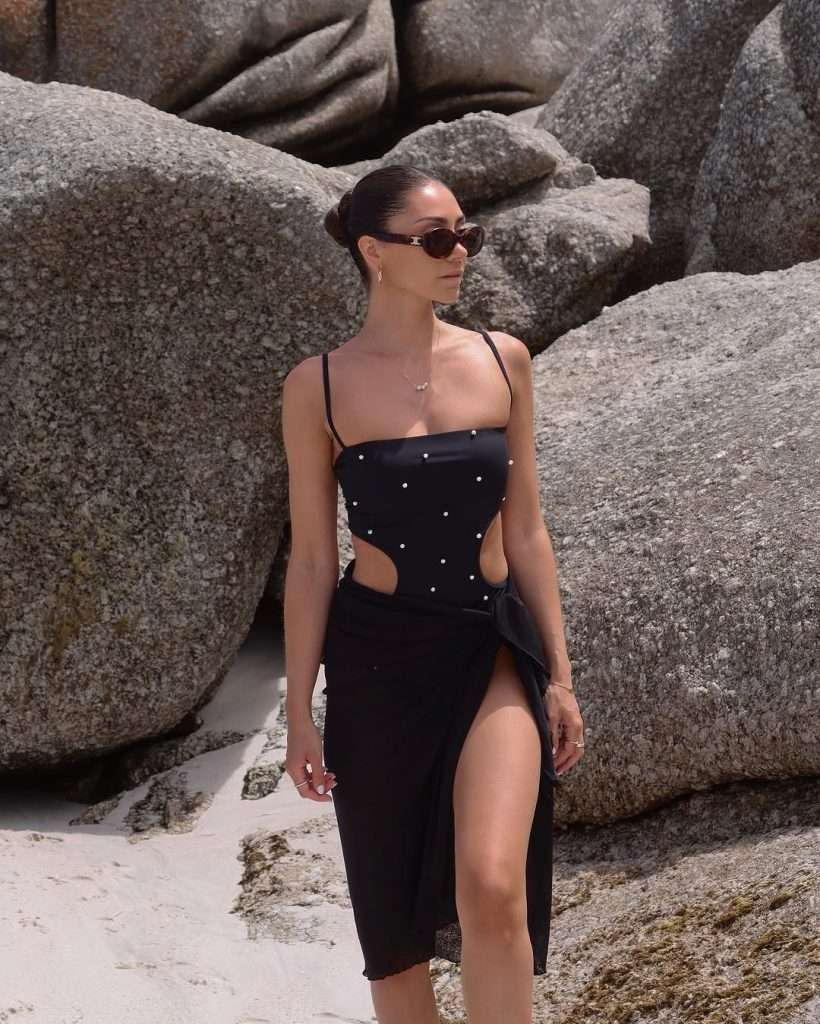 Beach vacation outfit with black one-piece swimsuit featuring pearl studs, side cutouts, matching black sarong skirt, dark sunglasses and delicate gold jewelry
