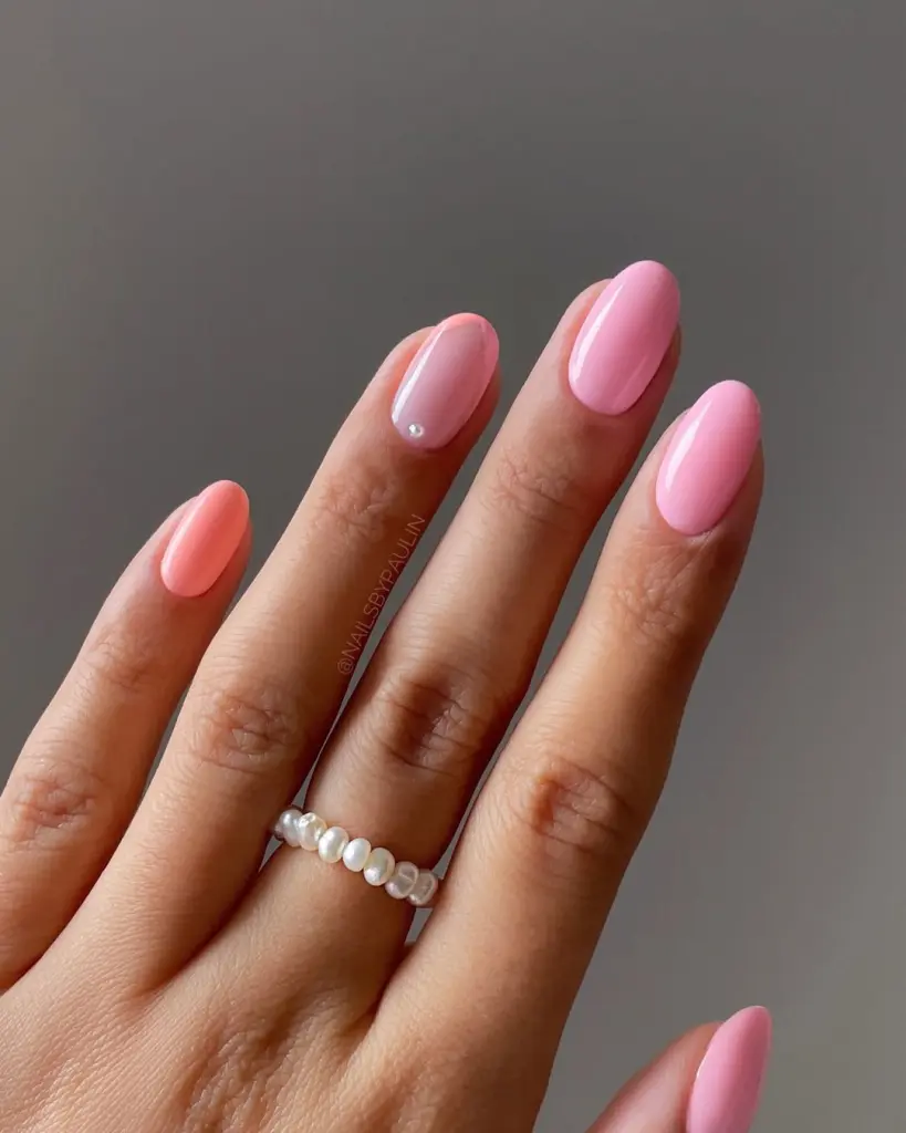 Short almond summer nails in varied pink shades from coral to light pink in skittle style with transparent nail featuring single pearl accent
