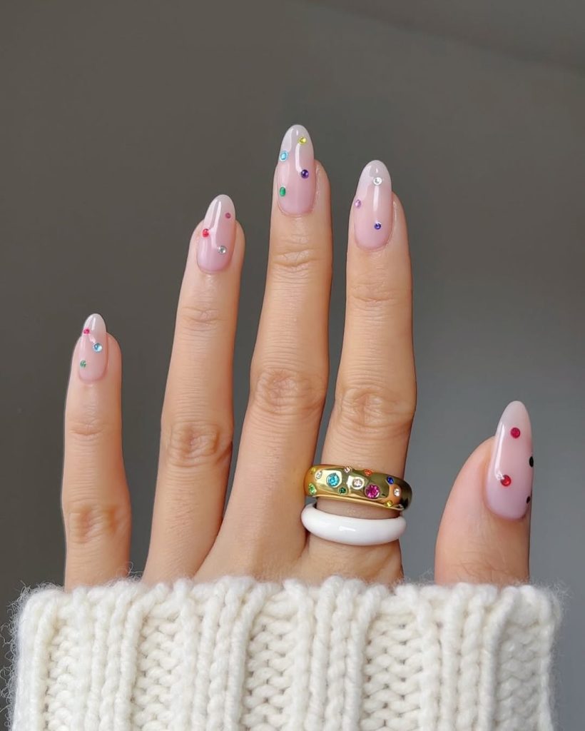 Almond shaped pink nails with scattered multicolored gemstone embellishments
