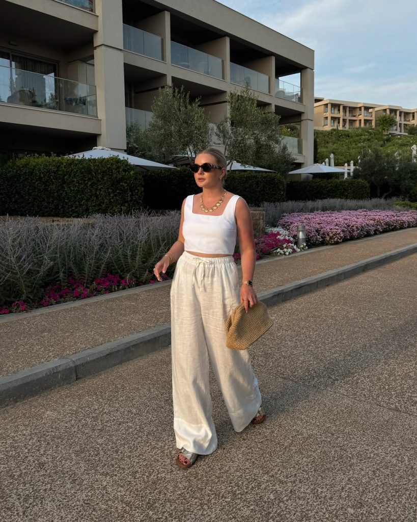 Elegant outfit with white square-neck crop top, cream wide-leg linen pants, gold chain necklace, straw clutch and oversized sunglasses
