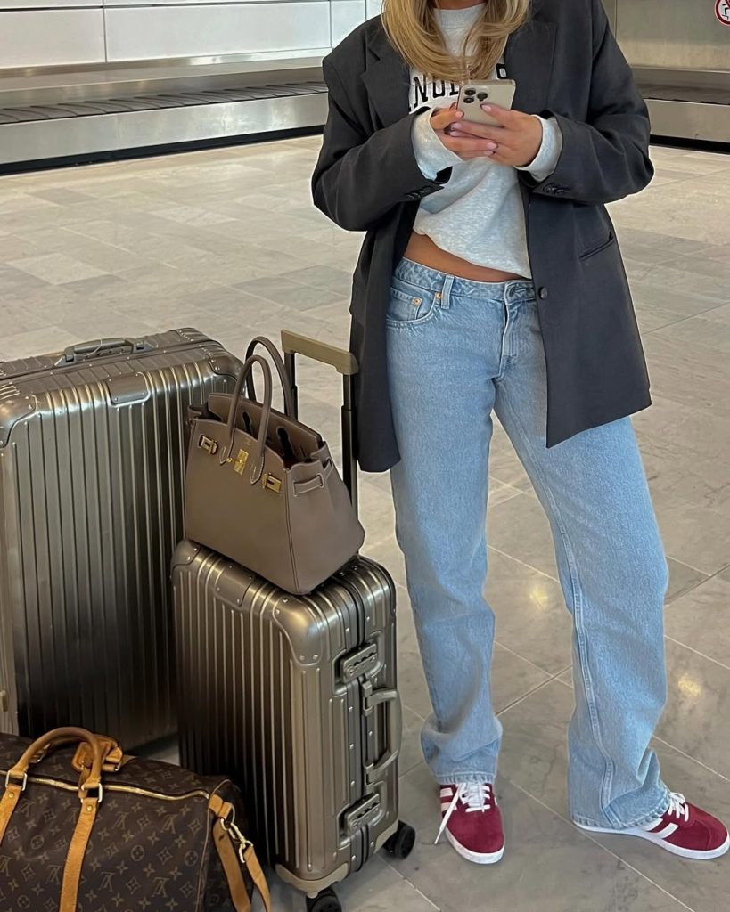 Travel outfit with oversized charcoal blazer, light gray graphic sweatshirt, light wash straight-leg jeans, burgundy sneakers and taupe designer handbag