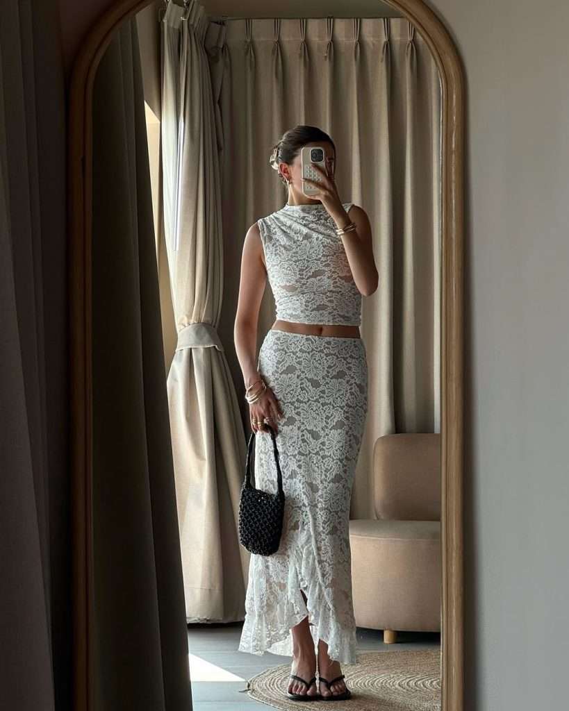 Beach vacation dinner outfit with white lace co-ord set featuring sleeveless crop top, high-waist maxi skirt with asymmetric ruffle hem, black beaded mini bag and flip flop sandals
