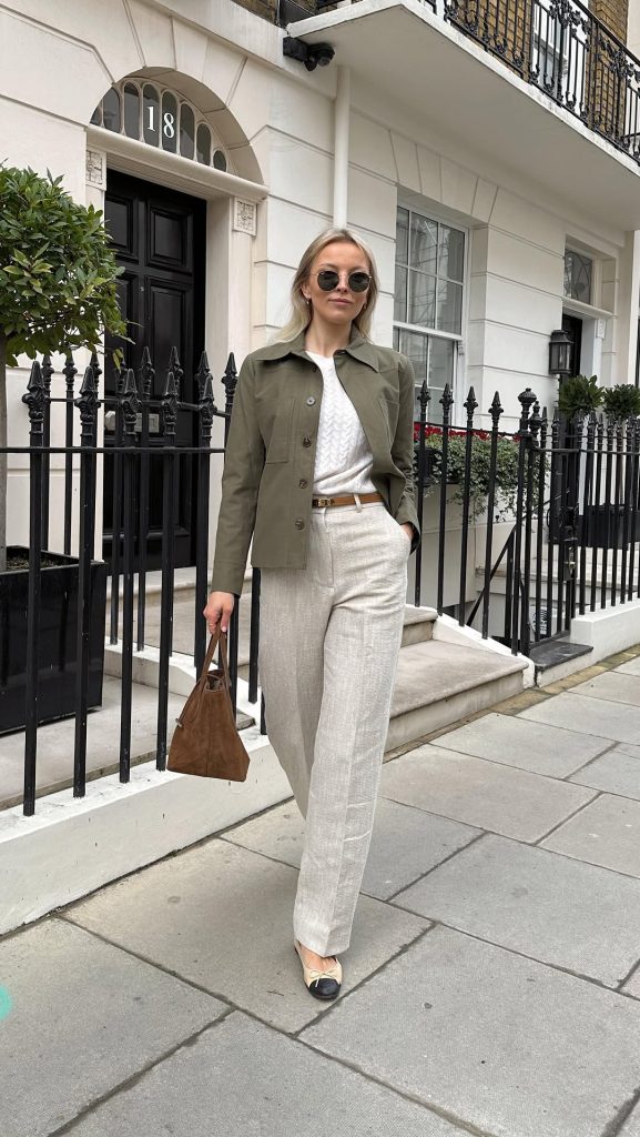 Elegant layered outfit with olive green utility jacket, white textured sweater, beige linen pants, tan leather belt, brown suede handbag and round sunglasses
