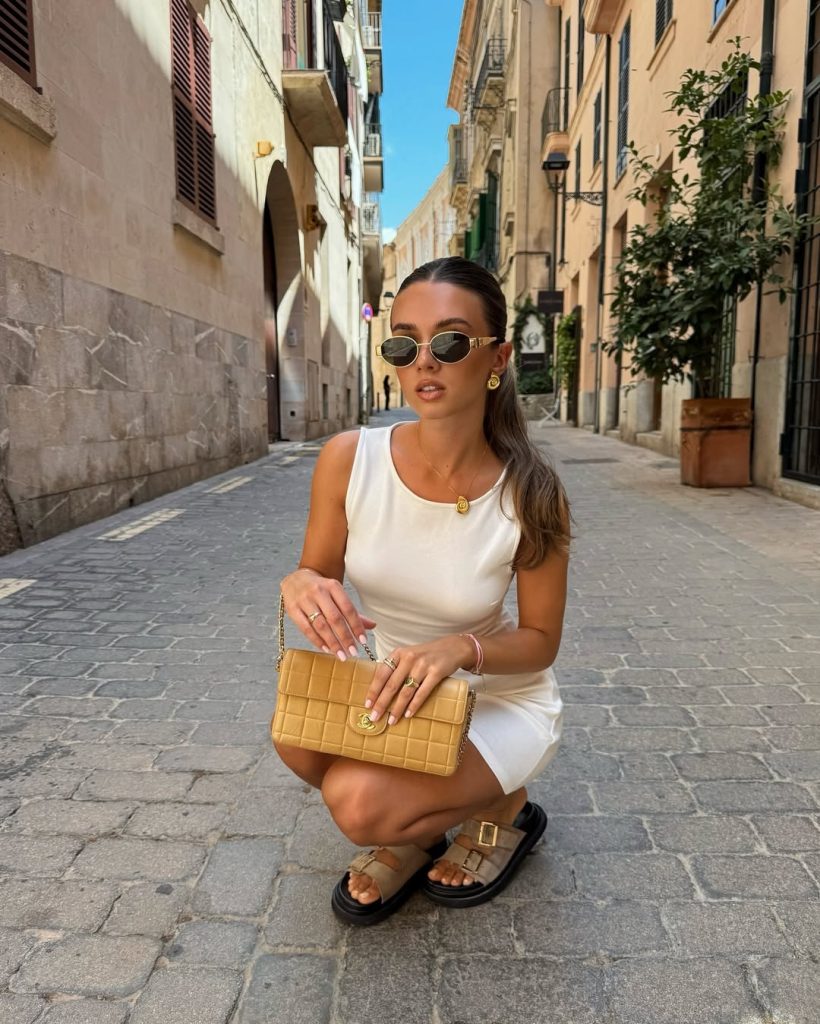 Elegant European vacation outfit with fitted white mini dress, beige quilted designer handbag, gold sunglasses and platform buckle sandals