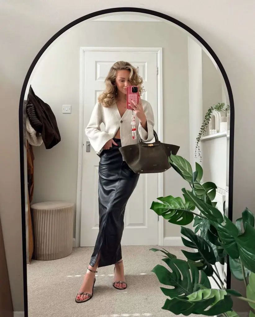 Chic spring date night outfit with cream cropped cardigan, black leather midi skirt, studded sandals and dark brown tote bag
