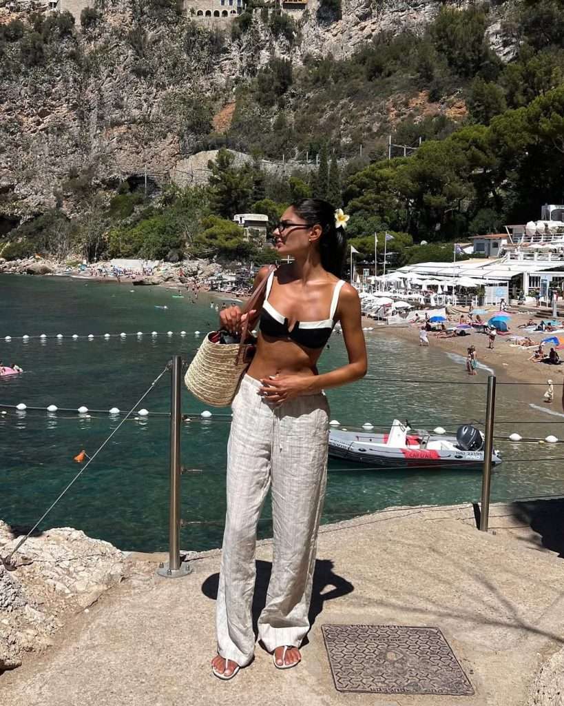 Beach vacation outfit with black and white contrast bikini top, beige linen pants, white flower hair accessory, straw bag and white sandals
