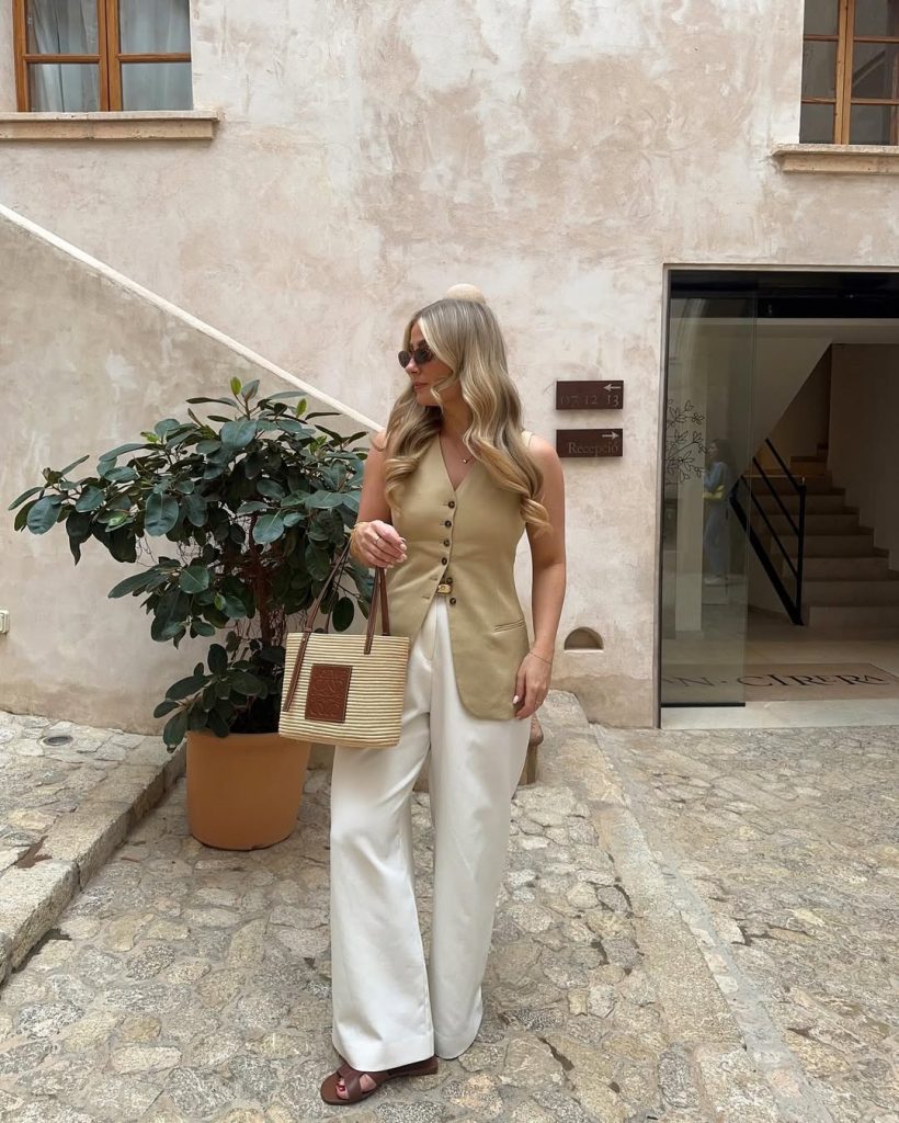 Elegant European vacation outfit with beige fitted button-front vest, white wide-leg trousers, striped canvas tote and brown leather sandals
