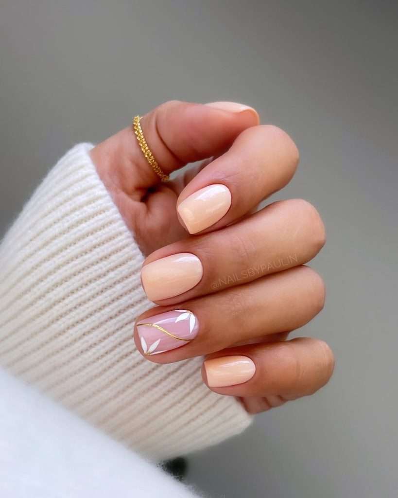 Short square nails in peach with milky accent nail featuring white leaf art and gold line detail
