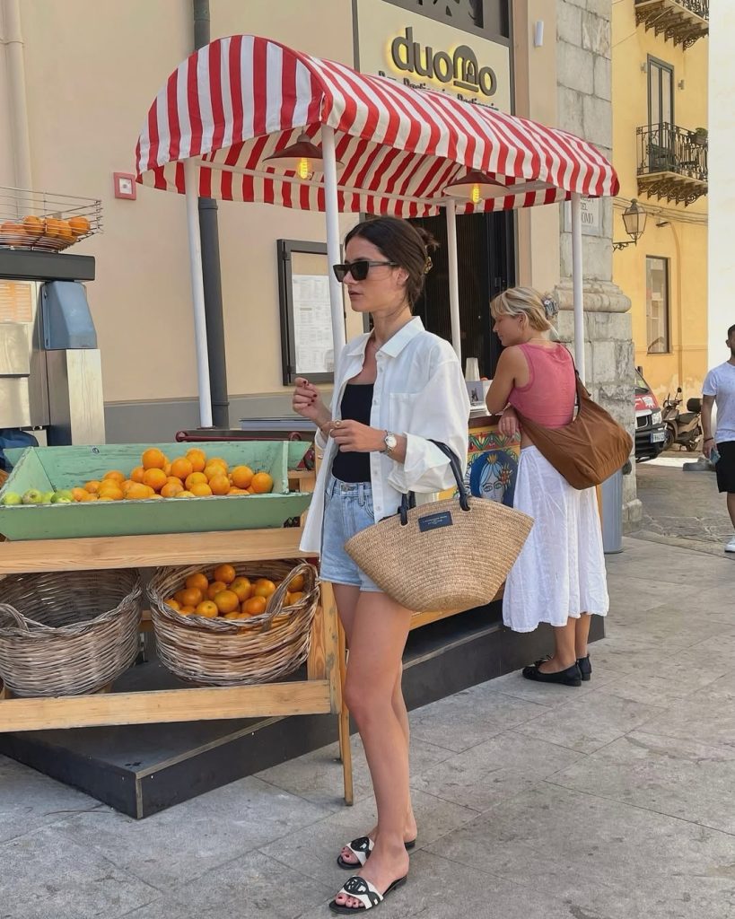  Casual European vacation outfit with white oversized button-down shirt, black tank top, light denim shorts, straw market bag and logo slide sandals 