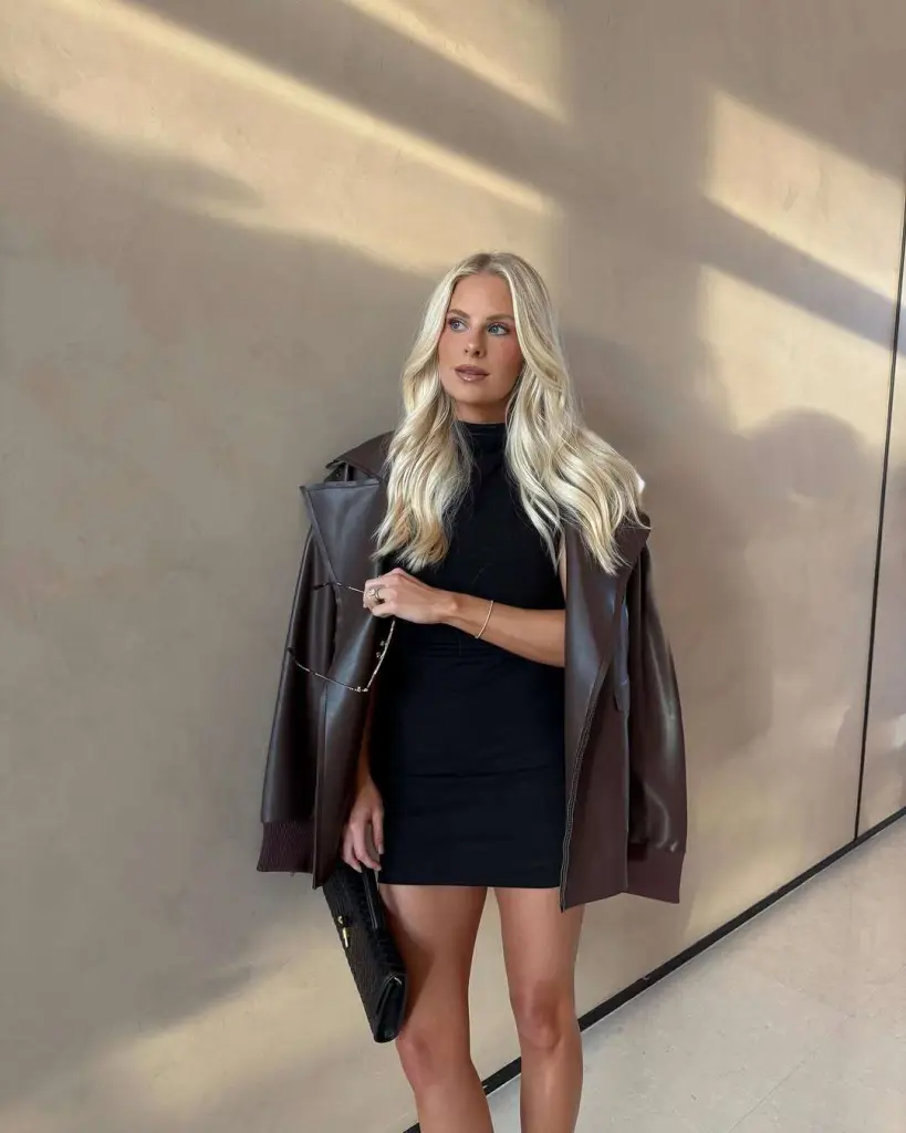 Stylish spring date night outfit with black mini dress, brown leather bomber jacket and black clutch purse