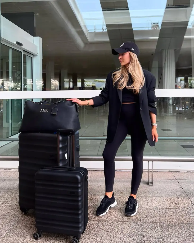 Chic airport outfit with black oversized blazer, black crop top, black leggings, navy baseball cap and black chunky sneakers