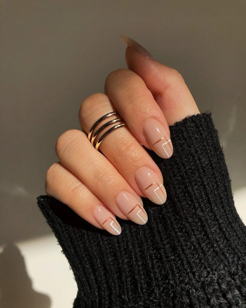 Almond-shaped minimalist nails with nude pink base featuring geometric white and gold linear designs across the nail beds