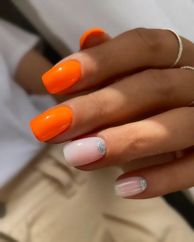 Short summer nails featuring bright neon orange polish on most fingers with accent nails in milky pink and silver glitter half-moon design