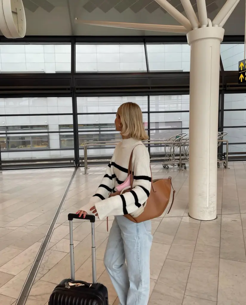 Stylish airport outfit with cream and black striped oversized sweater, light blue baggy jeans, tan leather tote bag and pink travel neck pillow