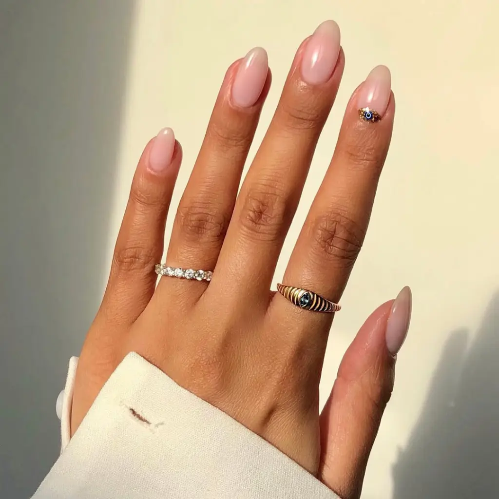 Almond-shaped sheer pink minimalist nails with single evil eye crystal charm surrounded by gold glitter on ring finger