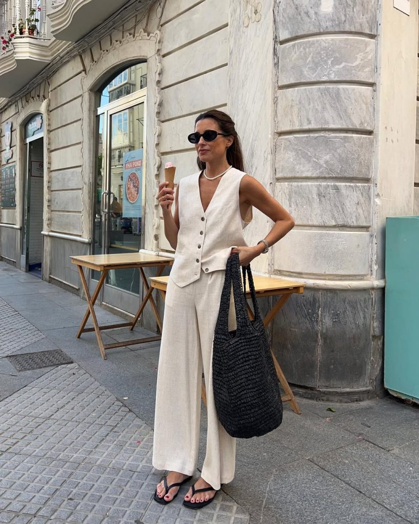 Chic European vacation outfit with cream linen button-front vest, wide-leg pants, black straw tote bag and minimal black sandals