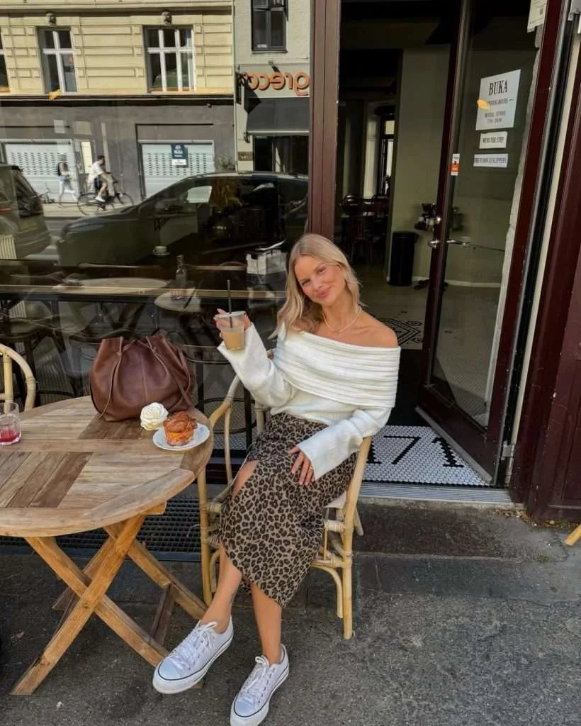 Casual spring date night outfit with cream off-shoulder sweater, leopard print midi skirt, white sneakers and brown leather bucket bag