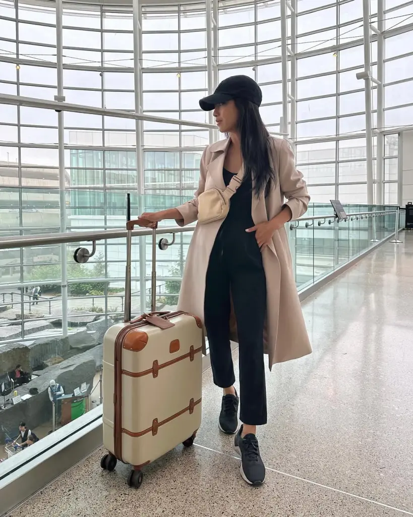 Stylish airport outfit with beige trench coat, black top, black trousers, black baseball cap, black sneakers and cream belt bag with vintage-inspired cream suitcase