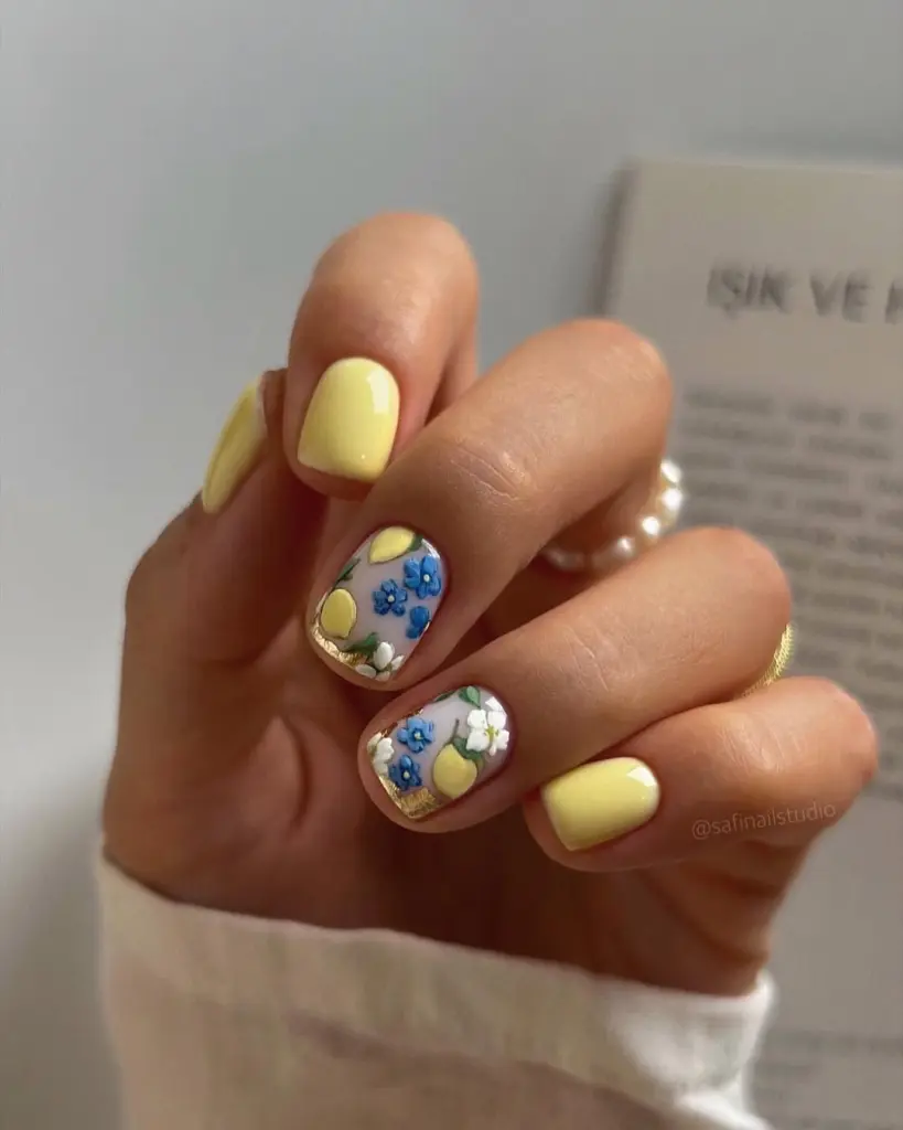 Short square summer nails with soft pastel yellow polish and accent nails featuring hand-painted blue and white flowers with lemon motifs on transparent base