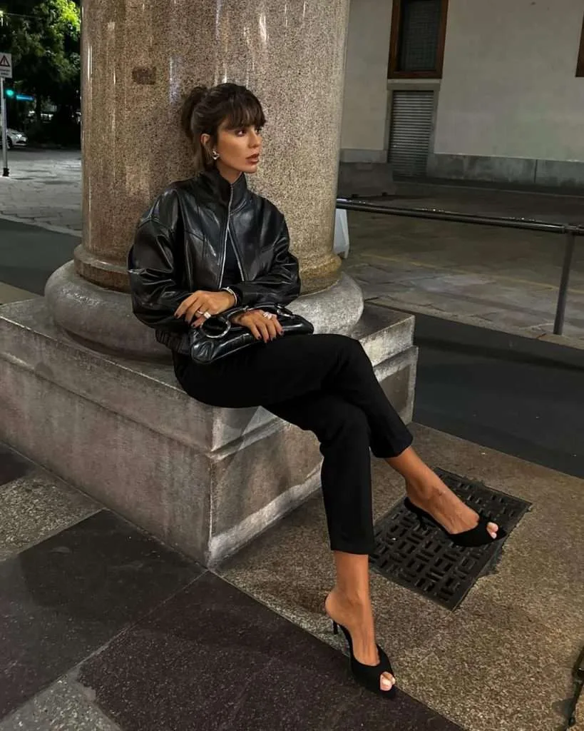Edgy spring date night outfit with black leather bomber jacket, black pants, black mule heels and matching clutch purse
