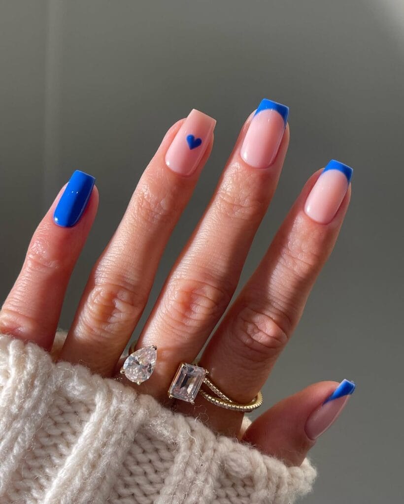 Square minimalist nails with nude pink base, cobalt blue French tips, blue accent nail, and small blue heart detail