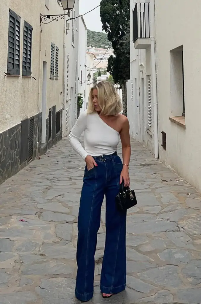 Chic summer date night outfit with white one-shoulder long sleeve top, dark wide-leg denim jeans, silver moon belt buckle and petite black handbag
