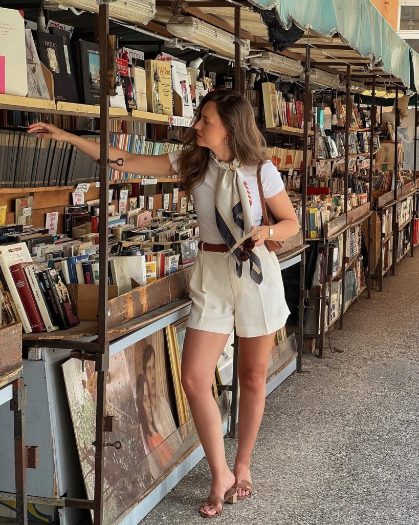 Old money summer outfit with white t-shirt, cream tailored shorts, silk neck scarf, brown leather belt, matching leather slide sandals and gold jewelry
